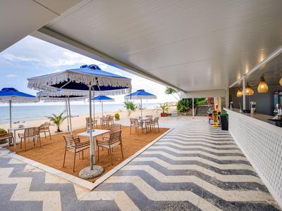 Offene Strandbar mit Sitzgelegenheiten unter weißen Sonnenschirmen und Blick auf das Meer.
