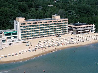 Grande hotel sulla spiaggia con ombrelloni e area boschiva adiacente.