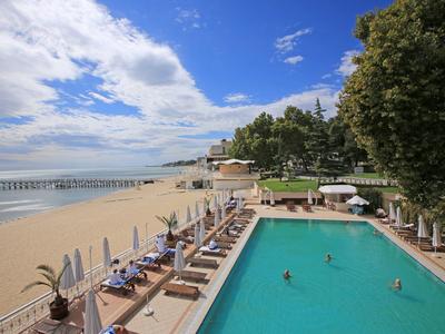 Piscina dell'hotel accanto a una spiaggia sabbiosa con lettini e alberi sotto un cielo azzurro.