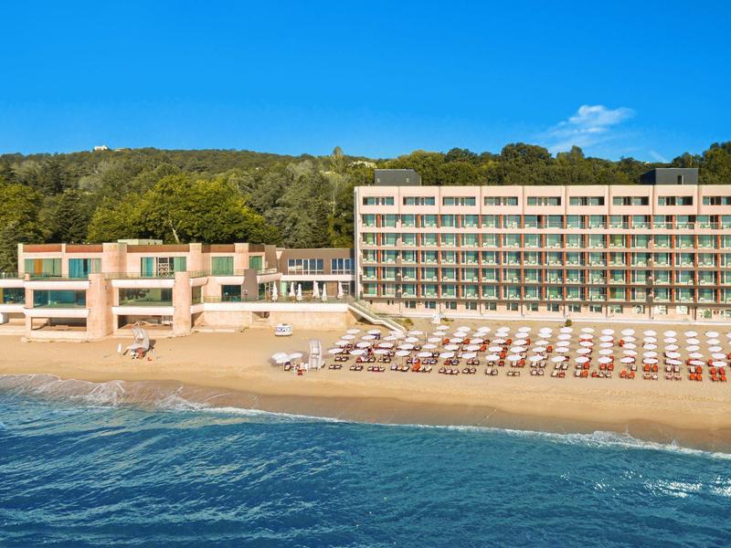 Hotel con terrazza, lettini e spiaggia sul mare davanti a collina boscosa
