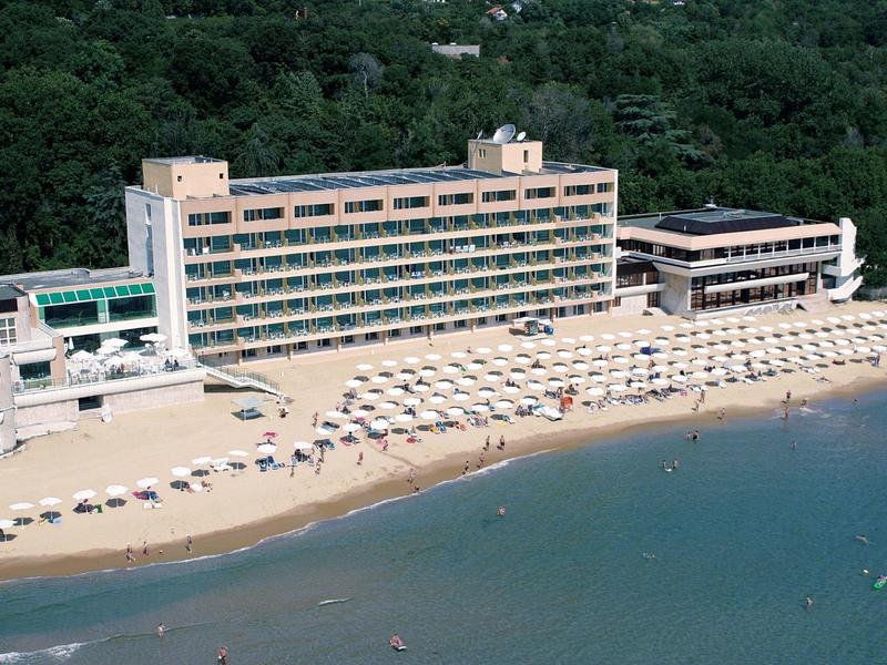 Grande hotel sulla spiaggia con ombrelloni e area boschiva adiacente.