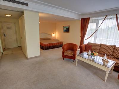 Bright hotel room with sleeping area, seating area, and large window with red curtains.