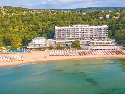 Large hotel on the beach with lounge chairs and wooded hills in the background.
