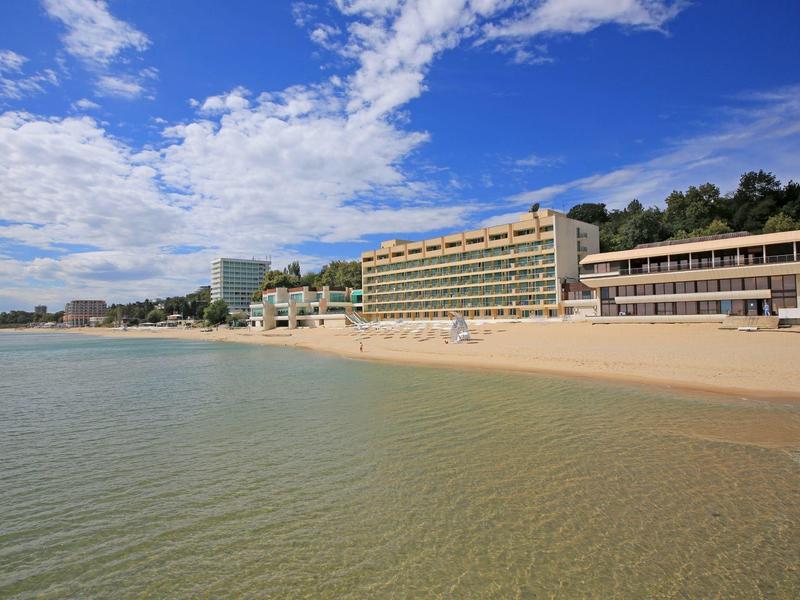 Beach with clear sea and large hotel building on the coast under a blue sky.