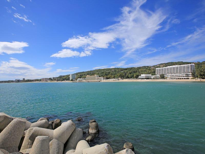 View of a beach with turquoise water, rocks in the foreground, and hotels on the horizon.