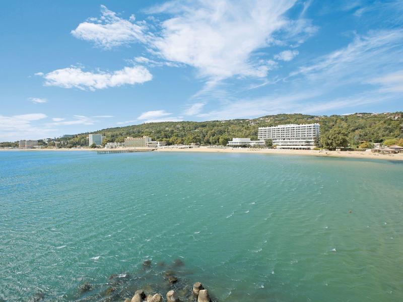 Coastline with clear water, sandy beach, wooded hills, and a hotel building in the background.