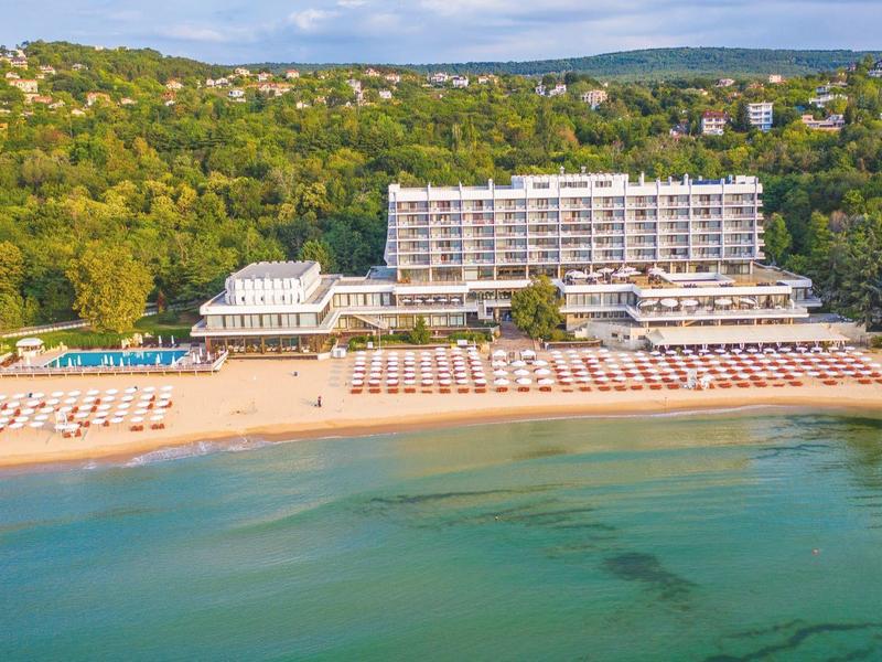 Large hotel on the beach with lounge chairs and wooded hills in the background.