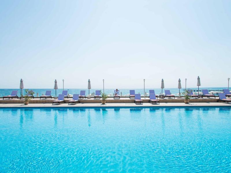Clear blue swimming pool with loungers and umbrellas on the beach in the background.
