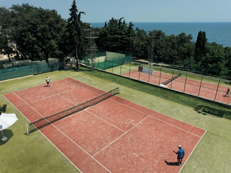 Two outdoor tennis courts next to the sea and trees with players on them.