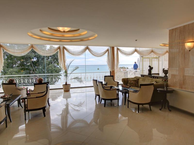 Elegant hotel lounge area with armchairs and tables, large glass front with sea view and curtains.