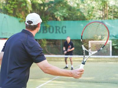 Twee mannen spelen tennis op een buitenbaan, één klaar om de bal te slaan.