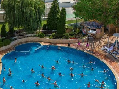 Druk zwembad in een hotelcomplex met uitzicht op de zee, omgeven door bomen en ligstoelen.