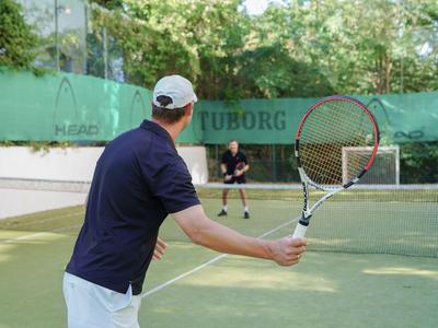 Due uomini giocano a tennis su un campo all'aperto circondato da una recinzione verde e alberi.