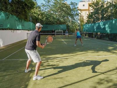 Due uomini giocano a tennis su un campo all'aperto soleggiato con recinzione verde e alberi.