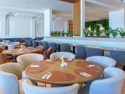 Area ristorante moderna dell'hotel con tavoli rotondi, sedie confortevoli e piante verdi come decorazione.
