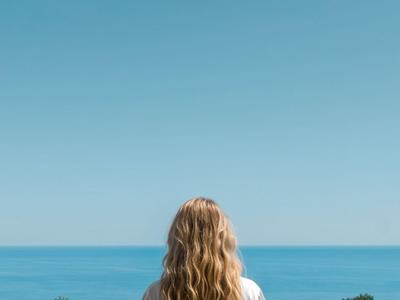 A woman with long blonde hair stands at a railing looking out at the sea.