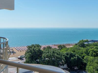 View from a hotel balcony overlooking the beach, sea, and green trees under a clear sky.