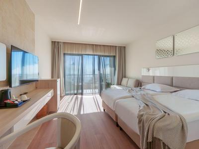 Bright hotel room with two beds, desk, chair, and view of balcony and sea.