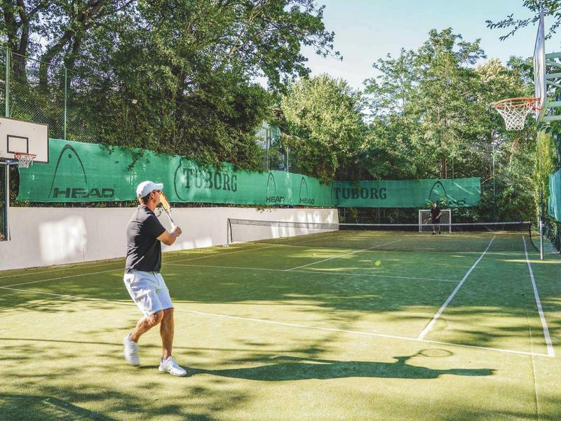 Un homme joue au tennis sur un court extérieur ensoleillé entouré d'arbres.