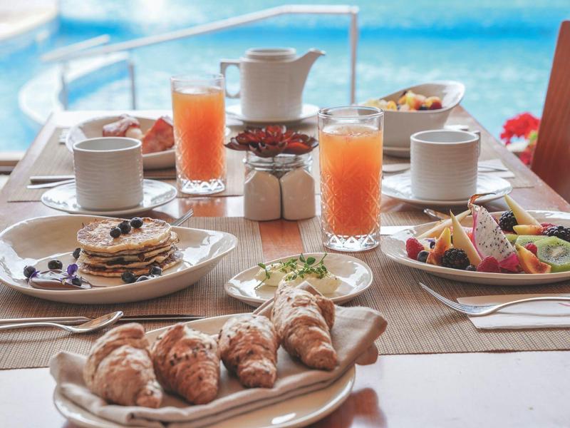 Table de petit déjeuner avec croissants, crêpes, jus et café près de la piscine en arrière-plan.