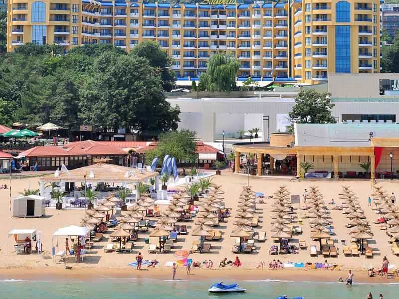 Grand hôtel avec de nombreux parasols et chaises longues sur une plage de sable devant une colline boisée.