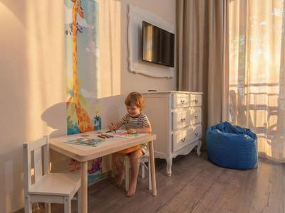 Kind zit aan tafel in lichte hotelkamer met tv en blauwe zitzak bij balkon.
