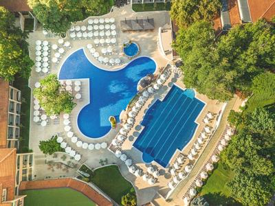 Luftaufnahme eines Hotelpools mit mehreren Formen, umgeben von weißen Liegestühlen und grünen Bäumen.