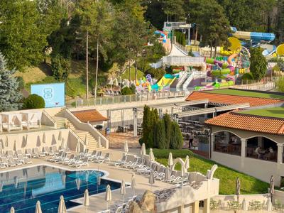 Hotel mit großem Außenpool, Liegestühlen und einem farbenfrohen Wasserspielplatz im Hintergrund.