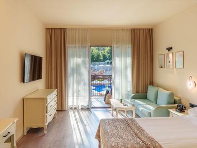 Helles Hotelzimmer mit Bett, Sitzbereich und Balkon mit Poolblick.