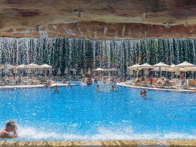 Großer Außenpool mit Sonne, Sonnenschirmen und umliegenden Bäumen in einem Urlaubsresort.