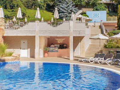Runder Pool mit Sonnenschirmen und Liegestühlen in einem Hotelgarten bei sonnigem Wetter.
