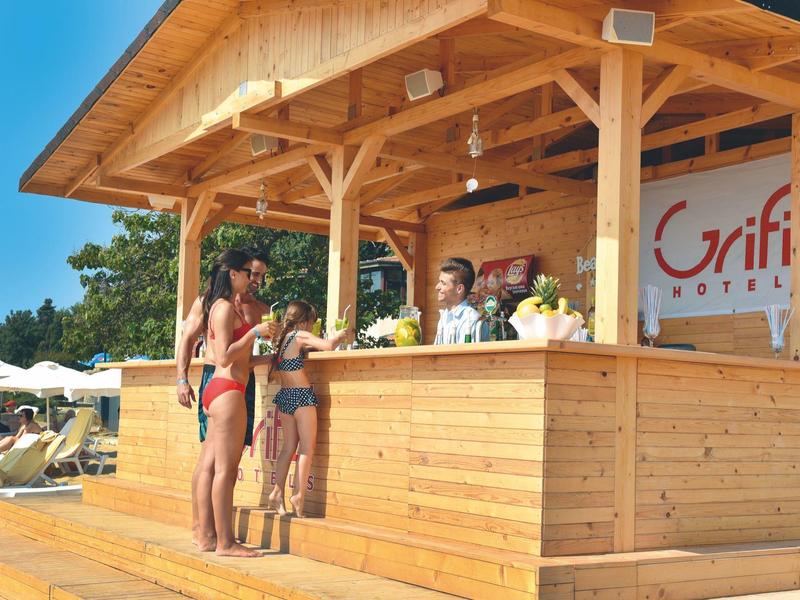 Een houten strandbar met gasten en barkeepers bij zonnig weer.