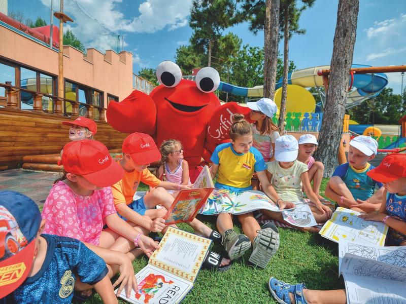 Kinderen zitten buiten op gras boeken te lezen, vergezeld door een persoon in een krabkostuum.
