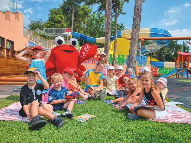 Kinderen zitten op het gras voor een waterpark met kleurrijke glijbanen en een krabmascotte.