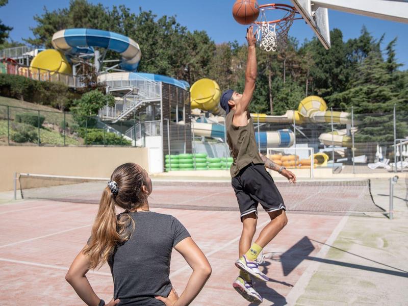 Twee mensen spelen basketbal op een veld met een waterpark en glijbanen op de achtergrond.