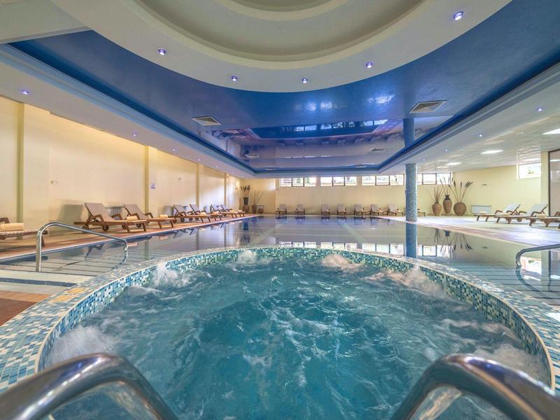 Binnenruimte van een hotel met jacuzzi op de voorgrond en zwembad op de achtergrond onder een blauwe plafond.