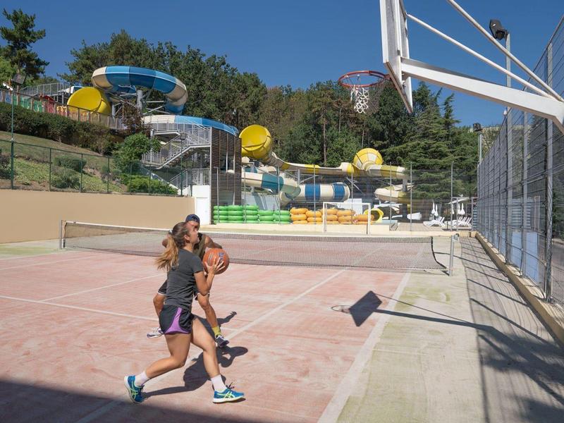 Twee mensen spelen basketbal op een buitenbaan naast een waterpark met glijbanen.