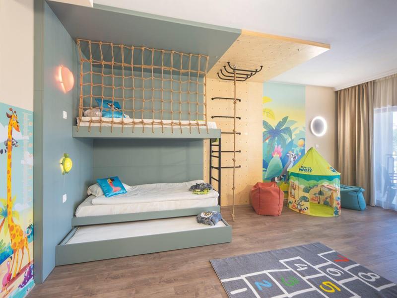 Kinderkamer met stapelbed, speelkleed en kleurrijke wanddecoratie in lichte kamer met raam.