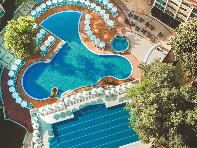 Luchtfoto van een hotel met meerdere zwembaden, parasols en ligstoelen omgeven door bomen.