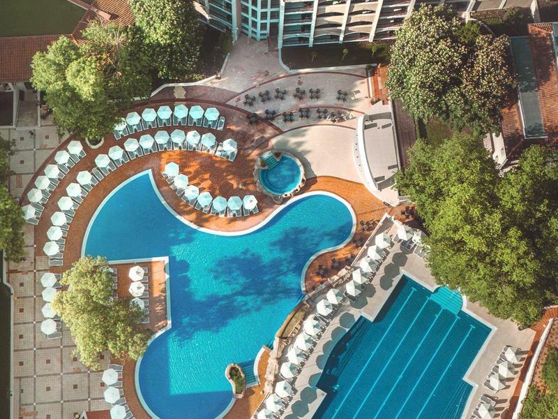 Luchtfoto van een hotel met twee grote zwembaden, parasols en omliggende bomen.