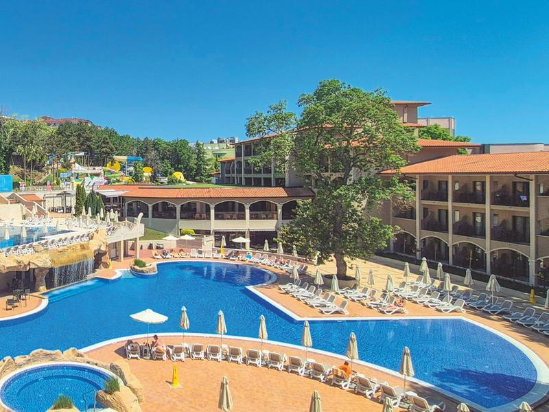 Hotel met groot buitenzwembad, zonneterrassen en omliggende begroeiing onder een blauwe lucht.