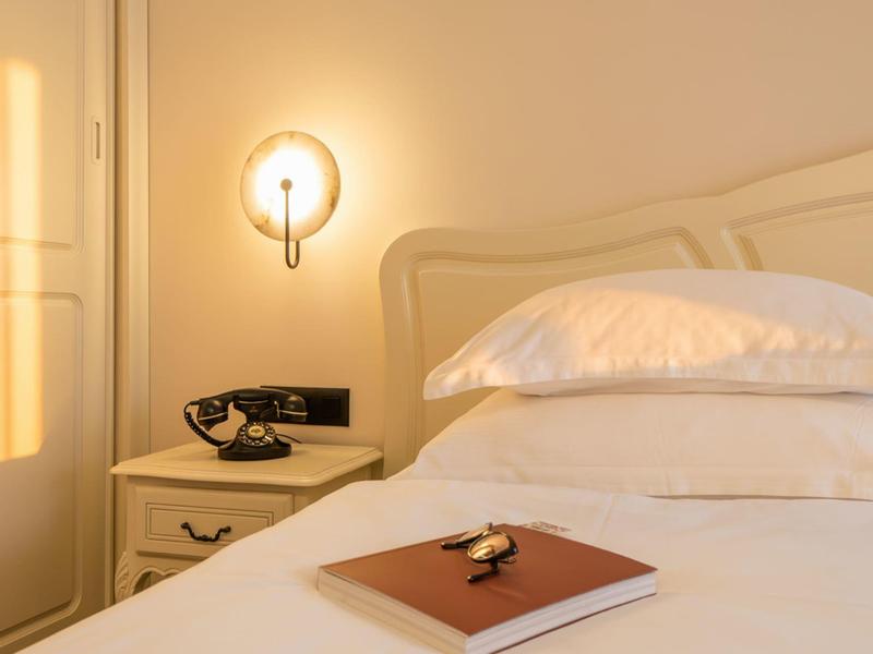 Gezellige hotelkamer met wit bed, nachtkastje, telefoon en wandlamp bij warm licht.