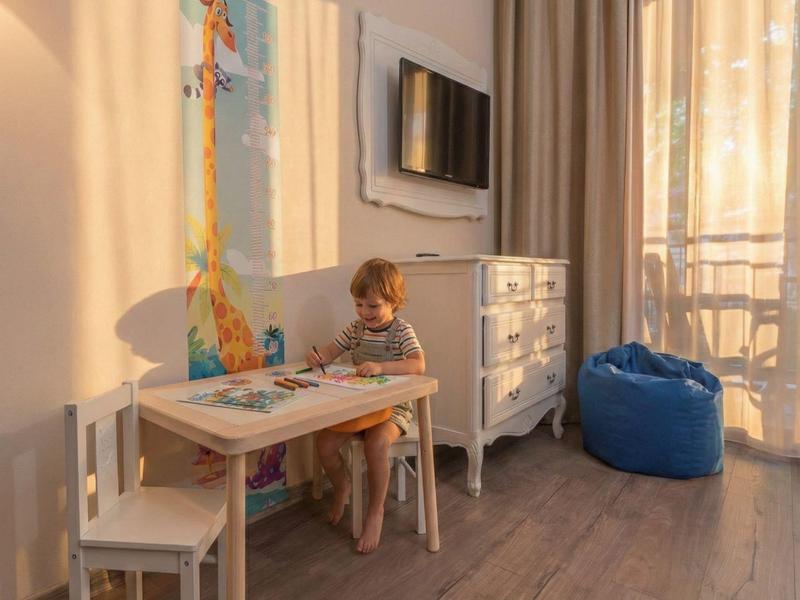 Kind zit aan tafel in lichte hotelkamer met tv en blauwe zitzak bij balkon.