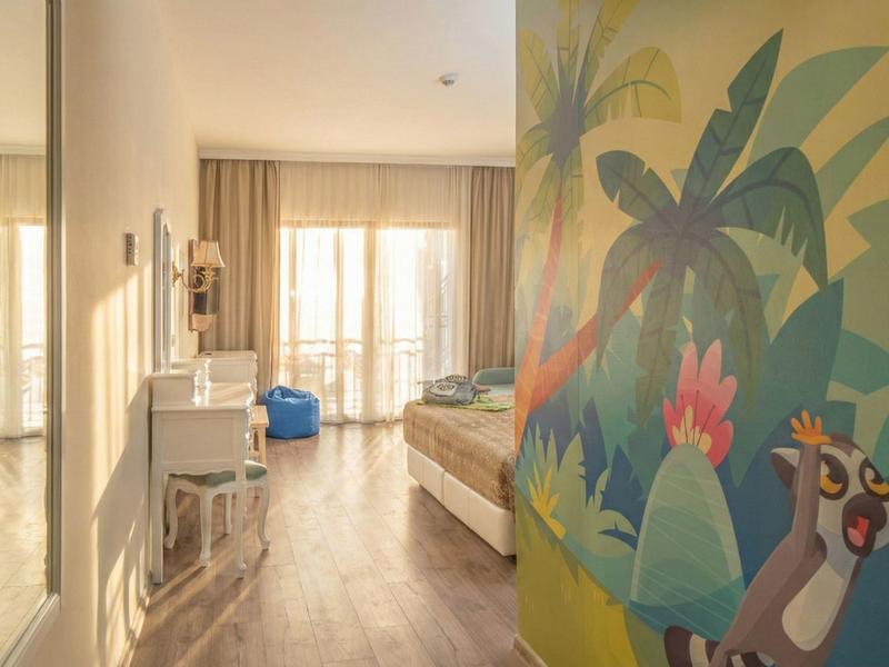 Lichte hotelkamer met houten vloer, groot bed en kleurrijke junglemuurschildering.