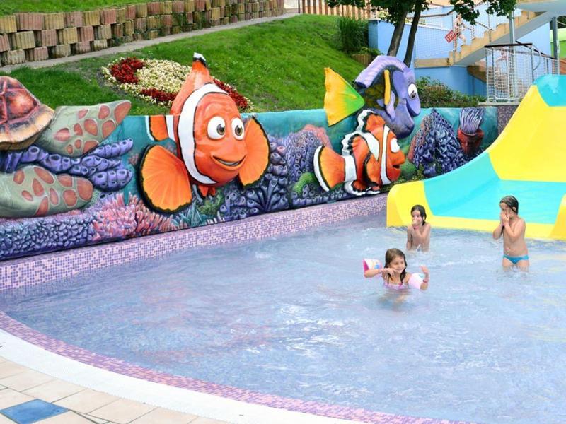 Kinderen spelen in een ondiep zwembad met een gele glijbaan en vissenmuurschildering in een resort.