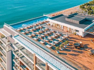 Moderne Dachterrasse mit Holzdeck, Pools, Sonnenliegen und Meerblick bei klarem Himmel.