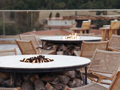 Outdoor-Terrasse mit runden Feuertischen, Holzlager und Flechtstühlen in natürlicher Umgebung.