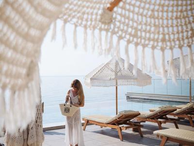 Frau in weißem Kleid unter Sonnenschirm an Strand mit Liegestühlen und Meerblick.