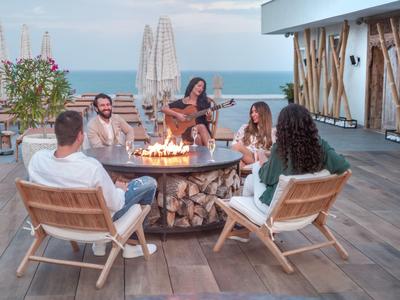 Five people sit around a circular fire pit on a terrace overlooking the sea.