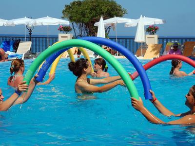 Menschen im Pool üben mit bunten Schaumstoffstäben unter blauem Himmel und weißen Sonnenschirmen.